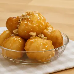 Des boules au miel dorées et croustillantes, garnies de noix concassées, servies dans un bol en verre sur une serviette à carreaux.