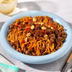 Un plat de nouilles au bœuf haché asiatique, garni de cacahuètes et de morceaux de poivrons rouges, servi dans une assiette bleue avec des baguettes.