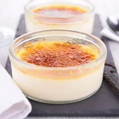 Deux crèmes brûlées verveine dans des ramequins en verre, avec une gousse de vanille et des couverts.