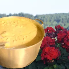 Un grand plat doré rempli d'une préparation crémeuse, évoquant la recette du patia, est posé à côté de fleurs rouges éclatantes, avec un paysage verdoyant en arrière-plan.