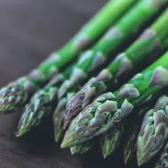 Un bouquet de pousses d'asperges vertes, aux pointes violettes, repose sur une surface en bois sombre.