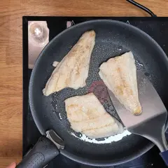 Trois filets de poisson blanc, parfaits pour une recette barracuda à la poêle, dorent dans une poêle sur une plaque de cuisson. Une spatule métallique soulève un morceau.