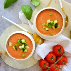 Deux bols de gaspacho melon concombre, garni de dés de concombre et de ciboule. Des tomates, un quartier de melon et des croûtons complètent la scène.