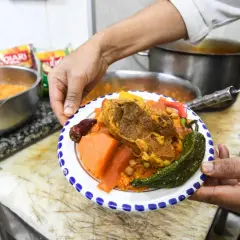 Une assiette de couscous garnie de viande, légumes et pois chiches, présentée par un cuisinier.