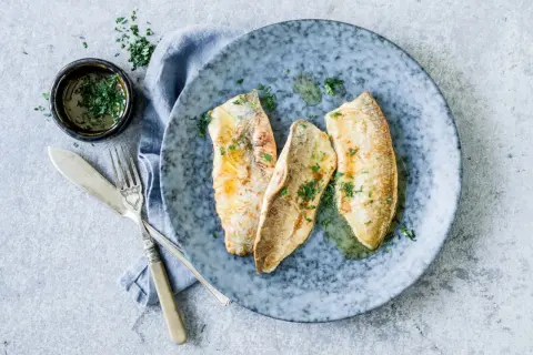 Trois filets de féra au four, parsemés de persil frais, sur une assiette bleue. Une petite coupelle de sauce et des couverts complètent cette recette simple.