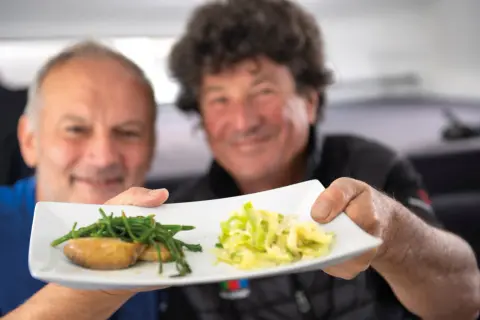 Deux hommes souriants présentent une assiette avec des ormeaux et des légumes. Une recette d'ormeau digne d'un grand chef.