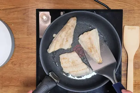 Trois filets de poisson blanc, parfaits pour une recette barracuda à la poêle, dorent dans une poêle sur une plaque de cuisson. Une spatule métallique soulève un morceau.