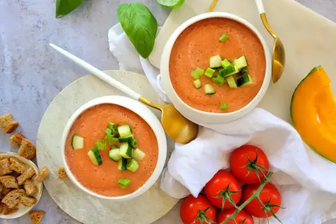Deux bols de gaspacho melon concombre, garni de dés de concombre et de ciboule. Des tomates, un quartier de melon et des croûtons complètent la scène.