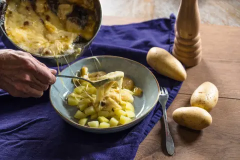 Une cuillère sert du fromage fondu sur des pommes de terre coupées, rappelant la saveur authentique de la matouille origine.