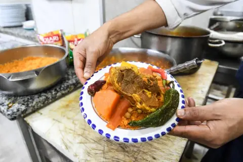 Une assiette de couscous garnie de viande, légumes et pois chiches, présentée par un cuisinier.