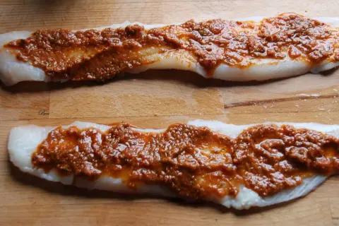 Deux filets de poisson blanc crus, recouverts d'une sauce rouge épicée, sur une planche à découper en bois.