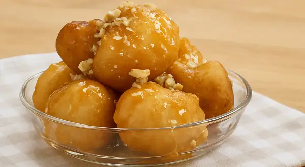 Des boules au miel dorées et croustillantes, garnies de noix concassées, servies dans un bol en verre sur une serviette à carreaux.