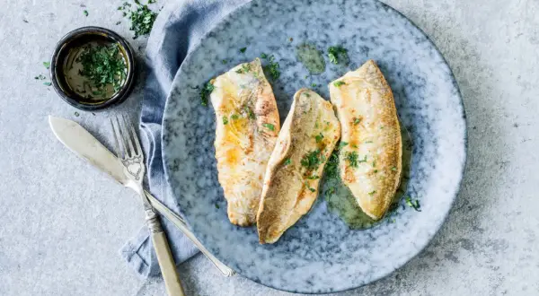 Trois filets de féra au four, parsemés de persil frais, sur une assiette bleue. Une petite coupelle de sauce et des couverts complètent cette recette simple.
