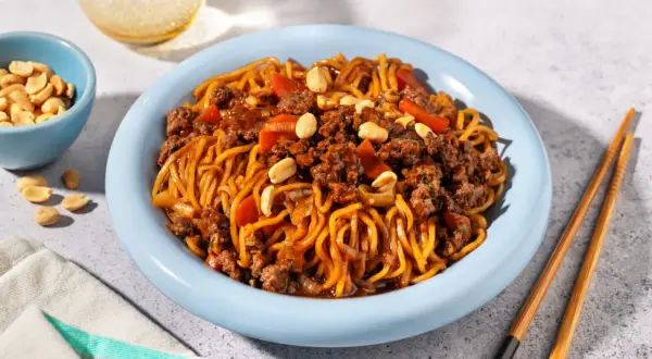 Un plat de nouilles au bœuf haché asiatique, garni de cacahuètes et de morceaux de poivrons rouges, servi dans une assiette bleue avec des baguettes.