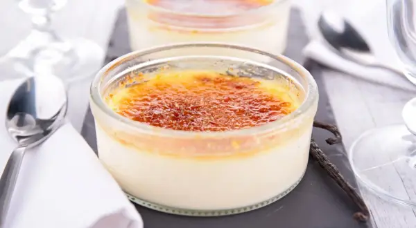Deux crèmes brûlées verveine dans des ramequins en verre, avec une gousse de vanille et des couverts.