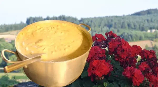 Un grand plat doré rempli d'une préparation crémeuse, évoquant la recette du patia, est posé à côté de fleurs rouges éclatantes, avec un paysage verdoyant en arrière-plan.