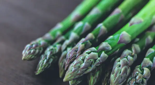 Un bouquet de pousses d'asperges vertes, aux pointes violettes, repose sur une surface en bois sombre.