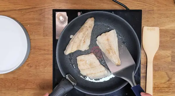 Trois filets de poisson blanc, parfaits pour une recette barracuda à la poêle, dorent dans une poêle sur une plaque de cuisson. Une spatule métallique soulève un morceau.