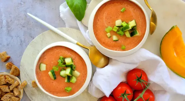 Deux bols de gaspacho melon concombre, garni de dés de concombre et de ciboule. Des tomates, un quartier de melon et des croûtons complètent la scène.