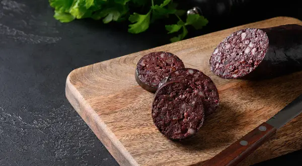 Tranché sur une planche en bois, un boudin noir froid révèle sa texture riche. Du persil frais et un moulin à poivre complètent la scène.