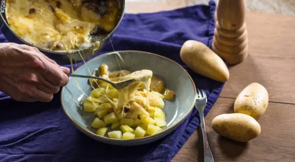 Une cuillère sert du fromage fondu sur des pommes de terre coupées, rappelant la saveur authentique de la matouille origine.