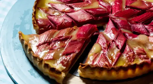 Tarte rhubarbe/pommes crémeuse aux éclats rouges et dorés, une part coupée sur une assiette bleue.