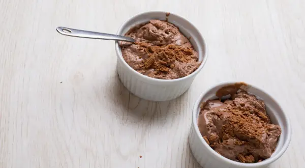 Deux ramequins de mousse au chocolat en poudre, l'un avec une cuillère, sur une table en bois clair.