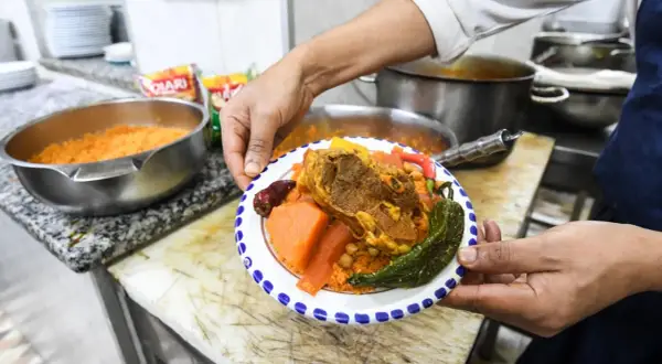 Une assiette de couscous garnie de viande, légumes et pois chiches, présentée par un cuisinier.