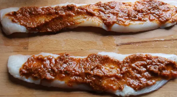 Deux filets de poisson blanc crus, recouverts d'une sauce rouge épicée, sur une planche à découper en bois.