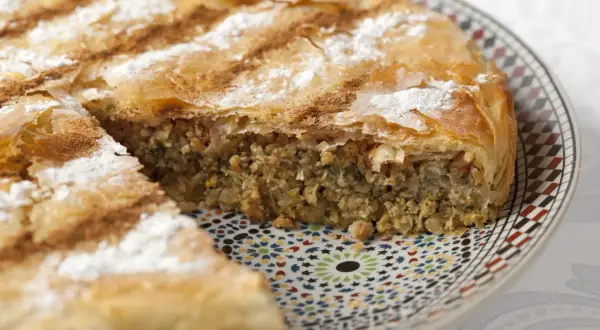 Une part de pastilla espagnol, feuilletée et saupoudrée de sucre glace et de cannelle, repose sur une assiette décorée.