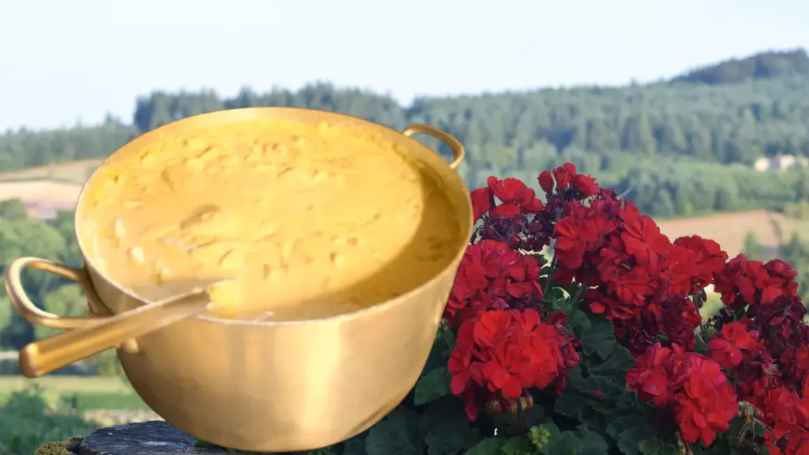 Un grand plat doré rempli d'une préparation crémeuse, évoquant la recette du patia, est posé à côté de fleurs rouges éclatantes, avec un paysage verdoyant en arrière-plan.