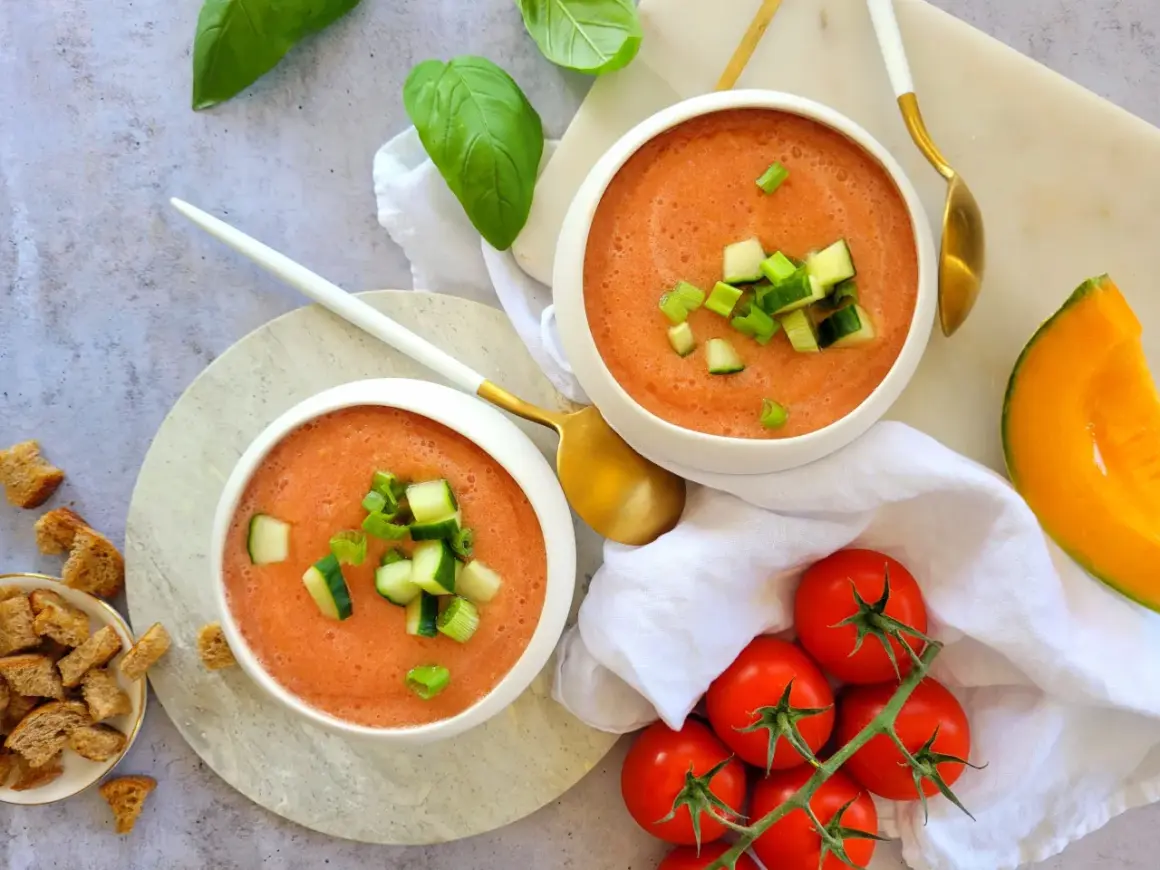 Deux bols de gaspacho melon concombre, garni de dés de concombre et de ciboule. Des tomates, un quartier de melon et des croûtons complètent la scène.