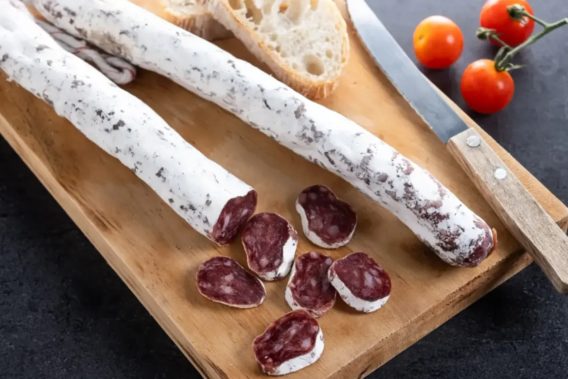 Deux saucissons secs, dont un tranché, sur une planche en bois avec du pain et des tomates cerises. Un délice de saucisson au genévrier.