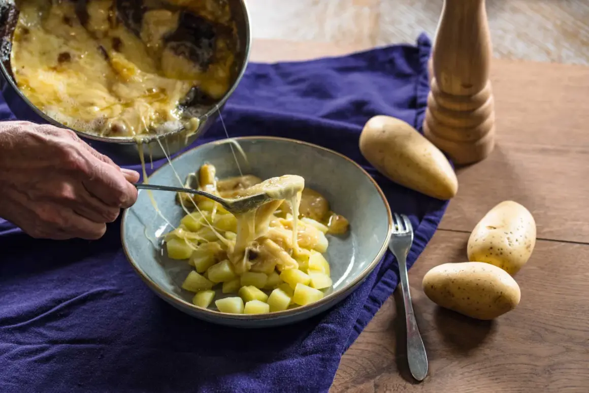 Une cuillère sert du fromage fondu sur des pommes de terre coupées, rappelant la saveur authentique de la matouille origine.