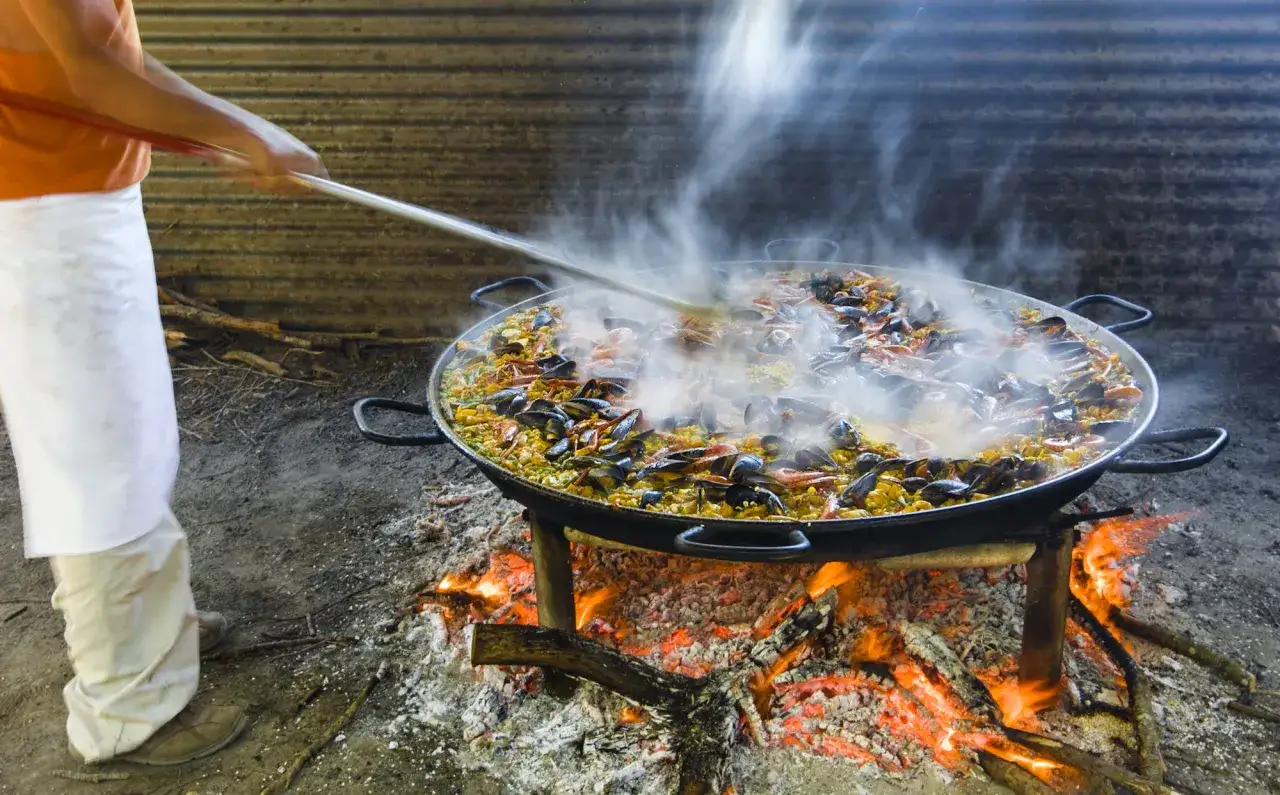 Une immense paella au feu de bois mijote, fumante et appétissante, avec des moules et des crevettes.