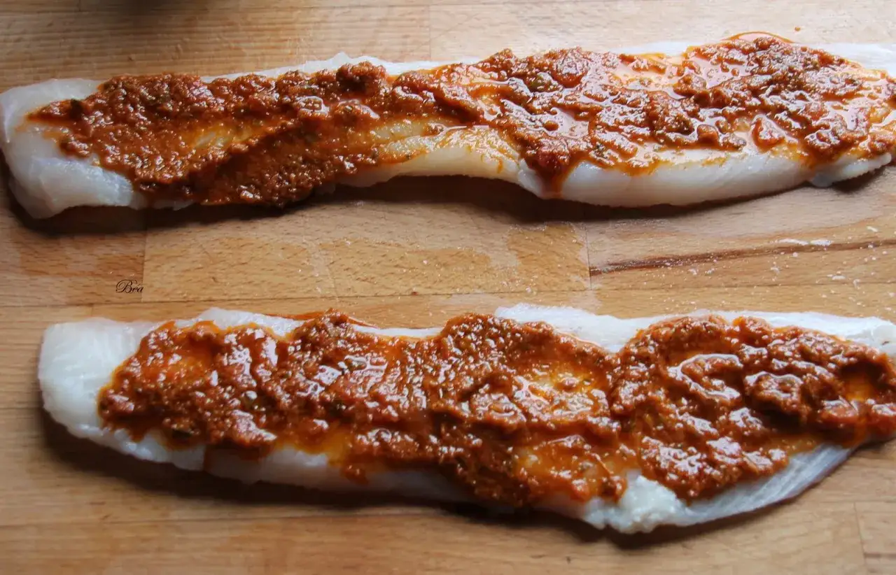 Deux filets de poisson blanc, probablement du filet de chimère, sont préparés sur une planche en bois, recouverts d'une sauce rouge épicée.