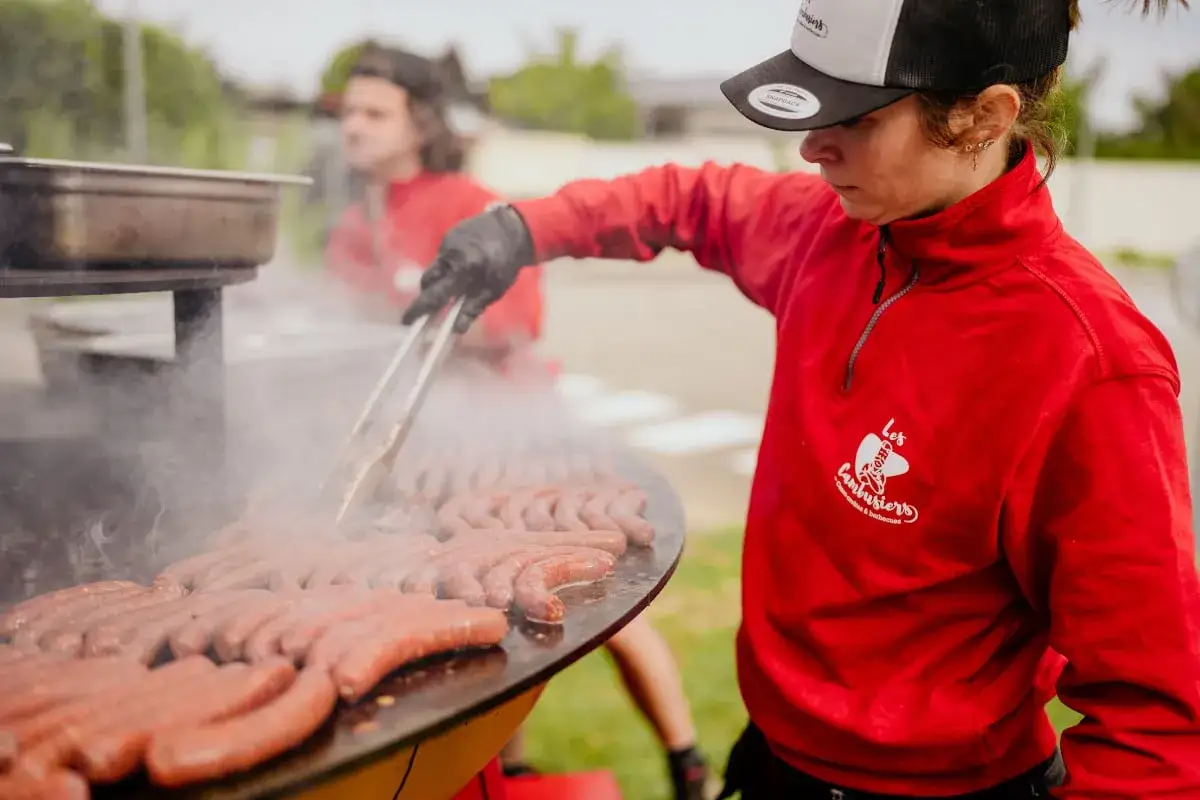 Une personne en sweat rouge prépare un gigot bitume sur un grand barbecue. La vapeur s'échappe des saucisses grillées.