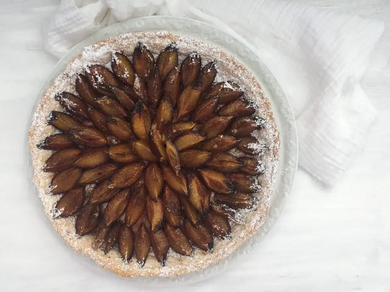 Tarte aux pruneaux et crème pâtissière, saupoudrée de sucre glace. Les fruits sont disposés en rosace sur une pâte dorée.