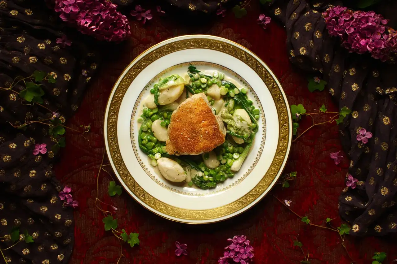 Un plat de ris de veau meunière, servi avec des petits pois et des pommes de terre, entouré de lilas et de tissu doré.