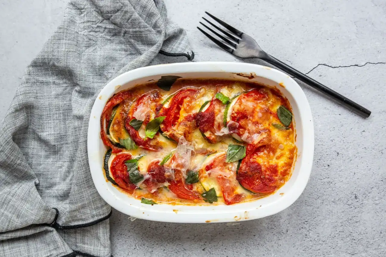 Un plat de gratin des Chartreux aux tomates et courgettes, gratiné au fromage, garni de feuilles de basilic frais.