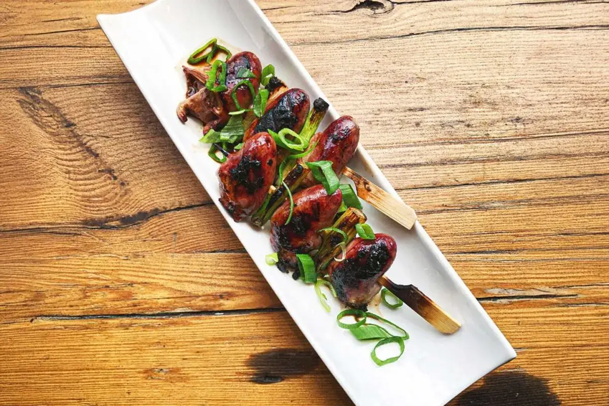 Brochettes de cœur de canard grillées, garnies d'oignons verts frais, sur une assiette blanche allongée.