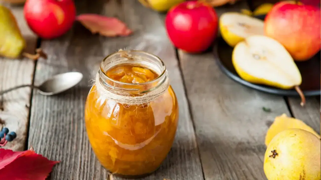 Un pot de compote pommes poires, orné de cordelette, sur une table en bois. Des pommes et poires fraîches, ainsi que des feuilles d'automne, complètent la scène.