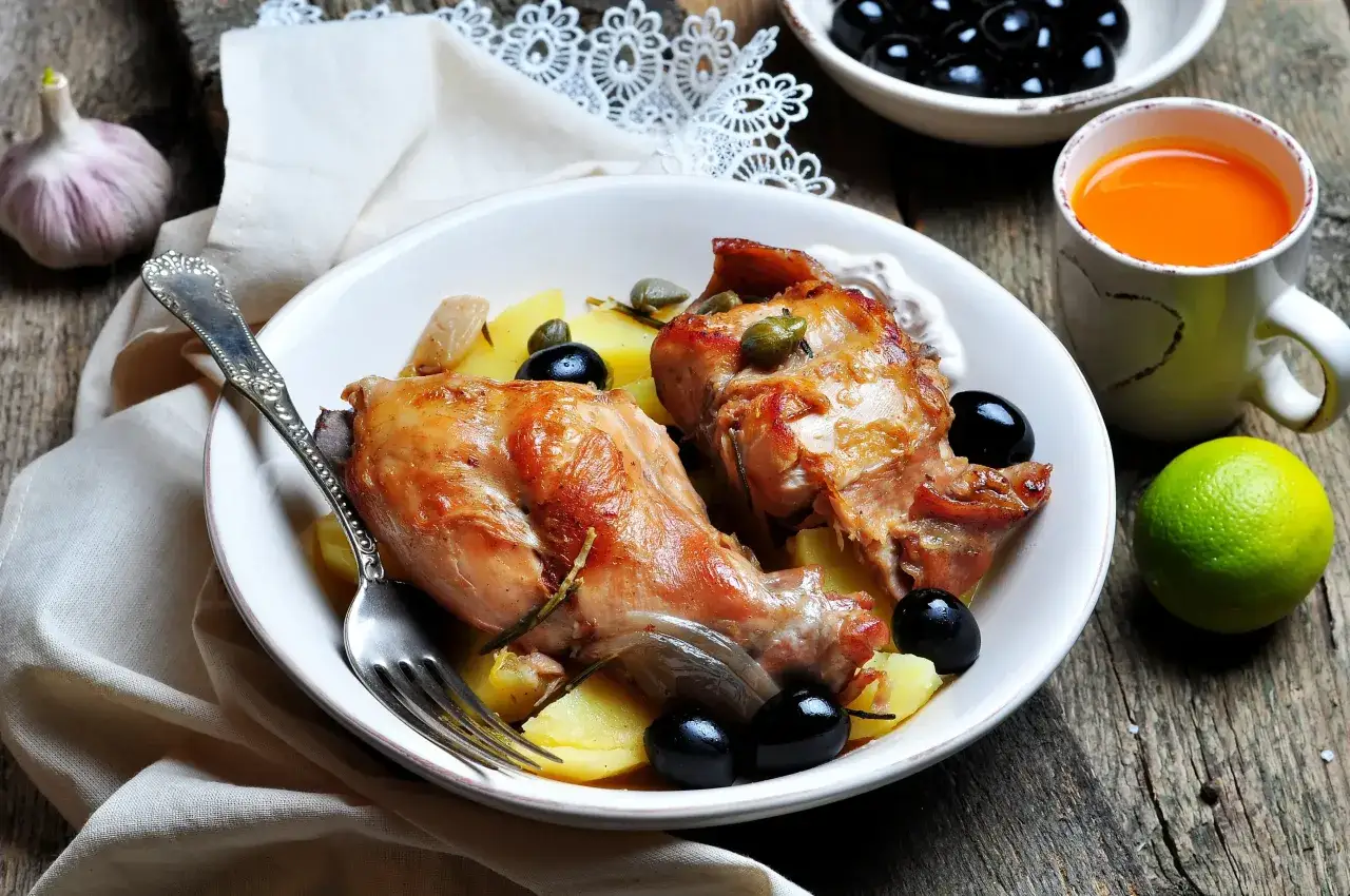 Caille sauce chasseur, plat rustique avec pommes de terre, olives noires et câpres. Une tasse de jus d'orange et une tête d'ail complètent la scène.