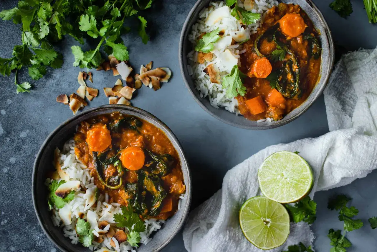 Deux bols de riz basmati servis avec un dahl de lentilles corail, carottes et épinards, garni de noix de coco grillée et de coriandre fraîche.