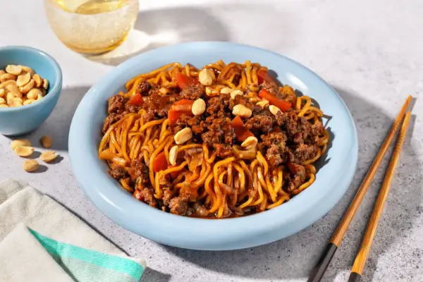 Un plat de nouilles au bœuf haché asiatique, garni de cacahuètes et de morceaux de poivrons rouges, servi dans une assiette bleue avec des baguettes.