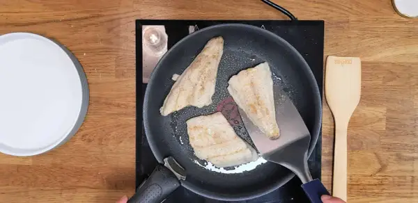 Trois filets de poisson blanc, parfaits pour une recette barracuda à la poêle, dorent dans une poêle sur une plaque de cuisson. Une spatule métallique soulève un morceau.
