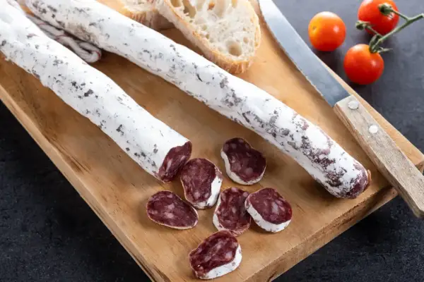 Deux saucissons secs, dont un tranché, sur une planche en bois avec du pain et des tomates cerises. Un délice de saucisson au genévrier.