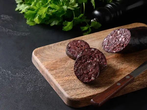 Tranché sur une planche en bois, un boudin noir froid révèle sa texture riche. Du persil frais et un moulin à poivre complètent la scène.