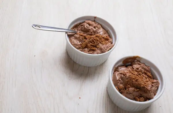 Deux ramequins de mousse au chocolat en poudre, l'un avec une cuillère, sur une table en bois clair.