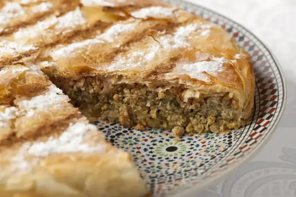 Une part de pastilla espagnol, feuilletée et saupoudrée de sucre glace et de cannelle, repose sur une assiette décorée.